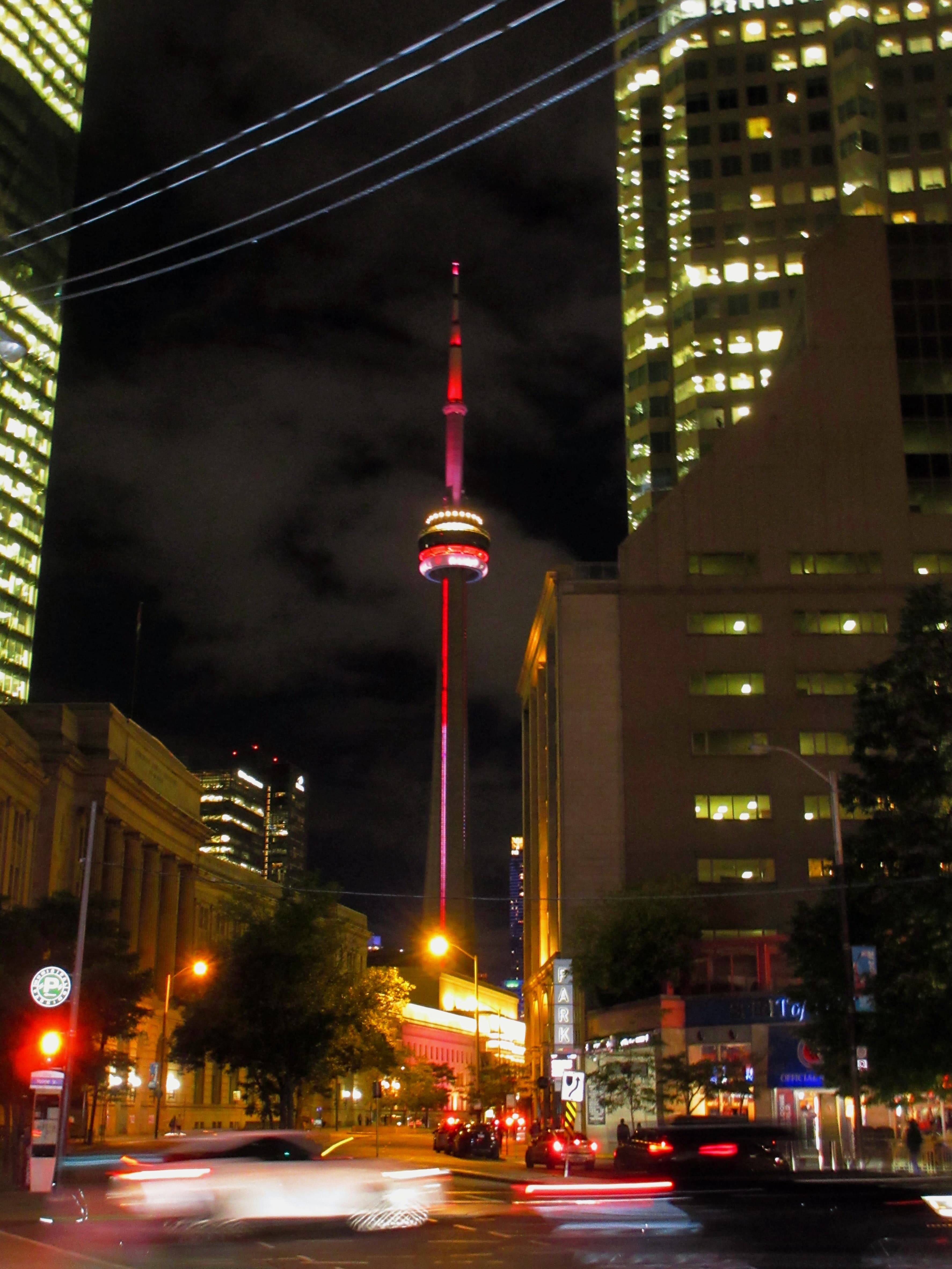 The CN Tower lit up at night between downtown Toronto office towers, with car lights streaking along the street below